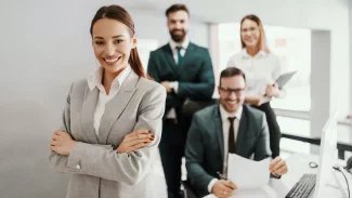 Usmívající se žena v obleku, za ní sedí a stojí dva muži a jedna žena.