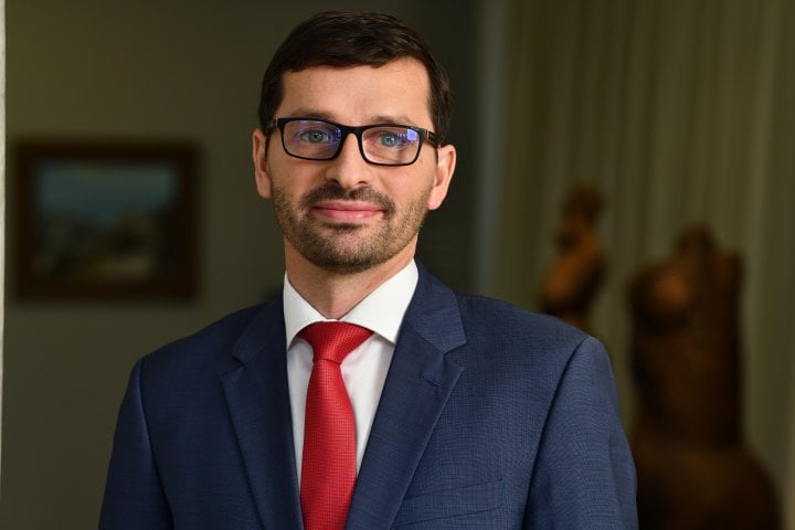 Muž v obleku s červenou kravatou a brýlemi.