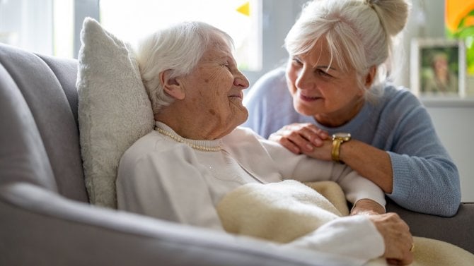 Starší žena s úsměvem pečující o nemohoucí seniorku.