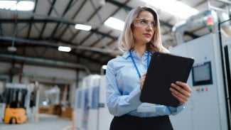 Žena v ochranných brýlích a s tabletem v ruce provádí kontrolu ve výrobní hale.