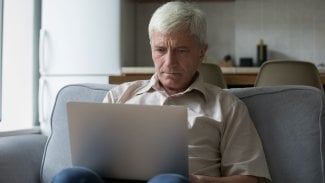 Straší muž sedí na gauči, má na kolenou notebook a kouká do něj. Tváří se zamyšleně
