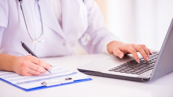 Detail rukou lékařky, která zapisuje do formuláře i počítače