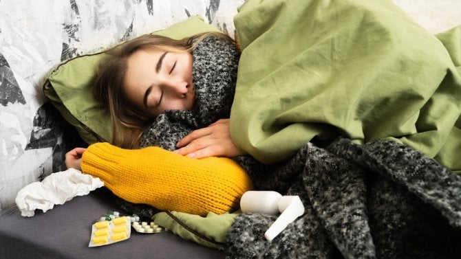 Nemocná žena leží se zimnicí v posteli, na nočním stolku má léky