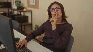 Žena sedící u stolu v kanceláři má prst na ústech (zákaz mluvení)