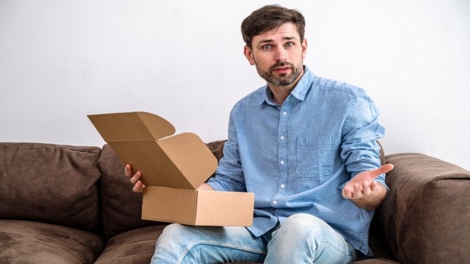 Muž sedí na pohovce a je rozčarován z rozbalování balíčku
