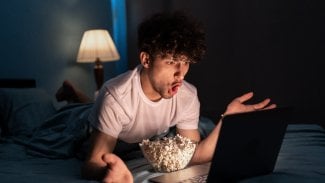 Mladý muž ležící v posteli se dívá do notebooku a před sebou má mísu s popcornem.
